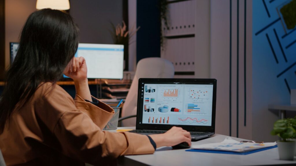 A person is analyzing data on a laptop with graphs and charts. Office environment with a modern aesthetic, blue accents, and desktop screens visible.