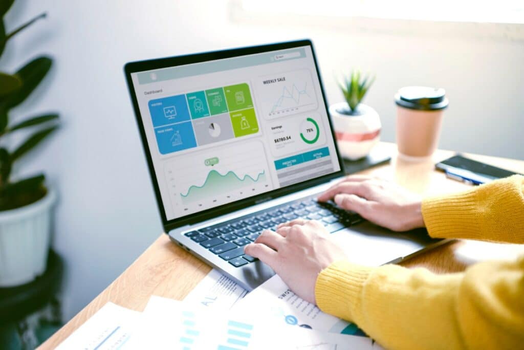 A person is analyzing financial data on a laptop with graphs displayed. Printed charts, a coffee cup, and houseplants are also on the desk.