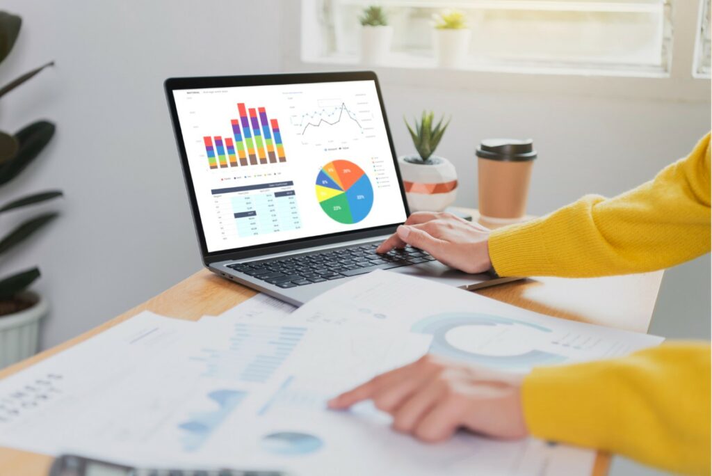 A person in a yellow sweater is analyzing graphs on a laptop with printed charts scattered on a desk, indicating data analysis or business work.