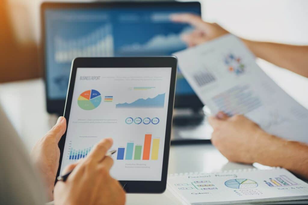 A person holds a tablet displaying colorful charts with another person pointing at a laptop screen, suggesting a business meeting with financial analysis.