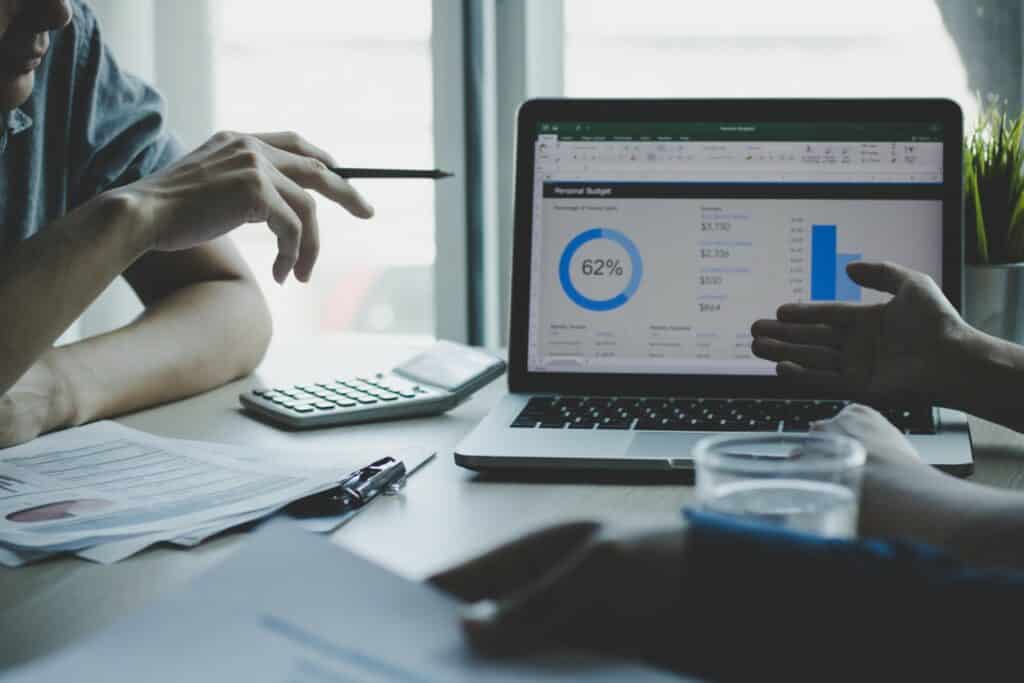 Two people are discussing finances with a pie chart on a laptop screen, papers, calculator, and water glass on a desk.