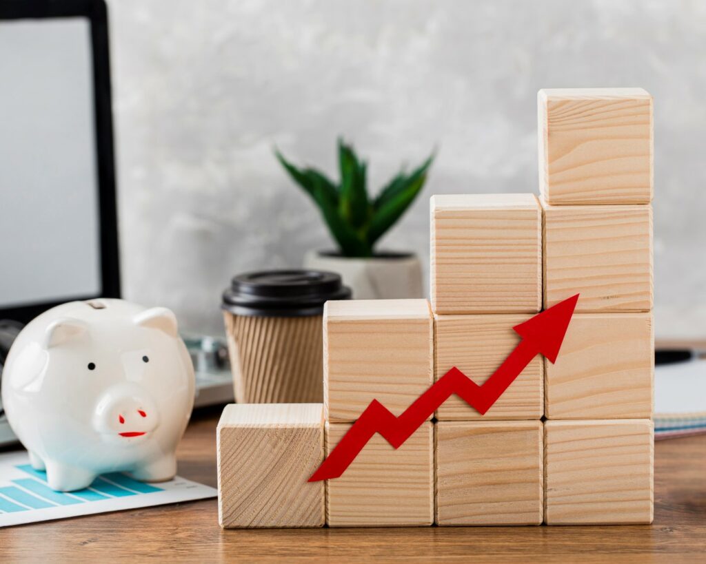 Wooden blocks arranged in a bar graph formation with a red upward arrow symbolize growth. A piggy bank, a coffee cup, and digital devices are in the background.