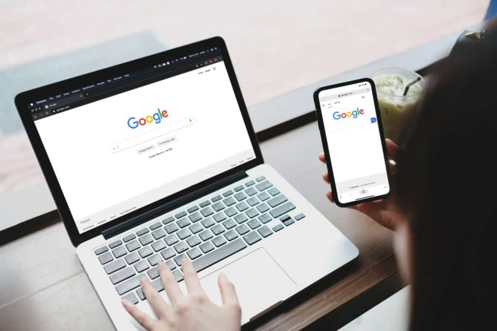 A person is using a laptop and a smartphone, both displaying the Google search page, on a desk with a glass of iced drink.