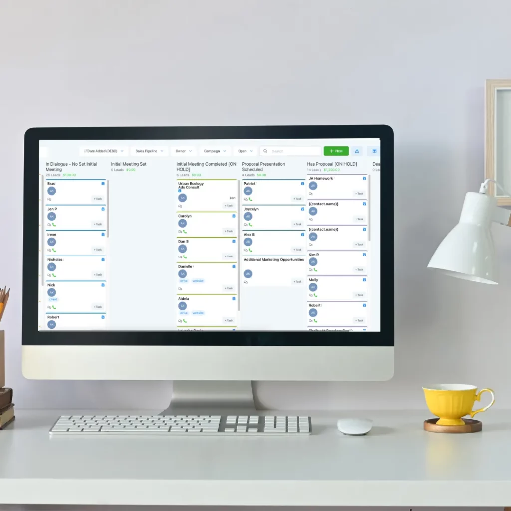 A neat workspace with an all-in-one computer displaying a sales pipeline application, alongside a white desk lamp, keyboard, mouse, and a yellow coffee cup.
