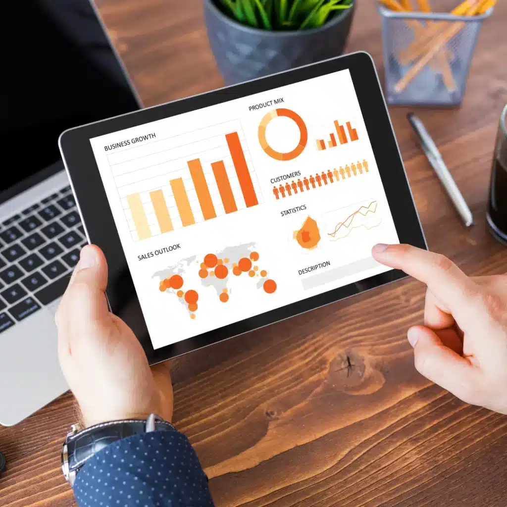 A person is holding a tablet displaying colorful business graphs and charts. A laptop, plant, pen, and pencils are also on the wooden desk.